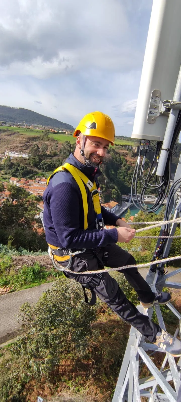 Técnico de Avanss con casco y arnés, trabajando en lo alto de una torre de antenas sobre un pueblo.